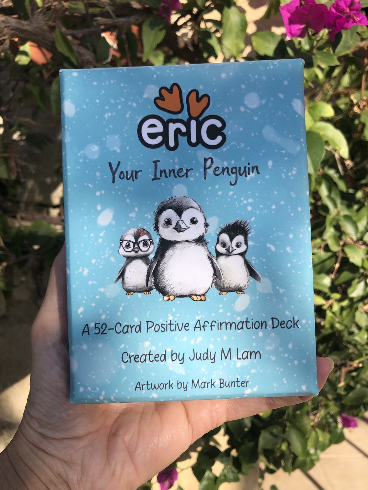 Your Inner Penguin affirmation cards: a 52-card positive affirmation deck held up among bougainvillea